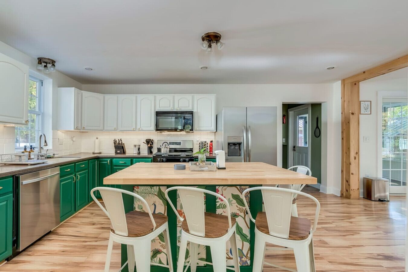 Una cocina moderna con gabinetes verdes, una isla central con sillas, electrodomésticos de acero inoxidable y un amplio ventanal que proporciona luz natural.