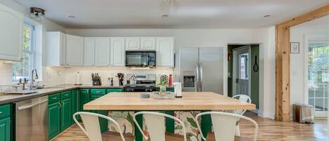 Una cocina moderna con gabinetes verdes, una isla central con sillas, electrodomésticos de acero inoxidable y un amplio ventanal que proporciona luz natural.