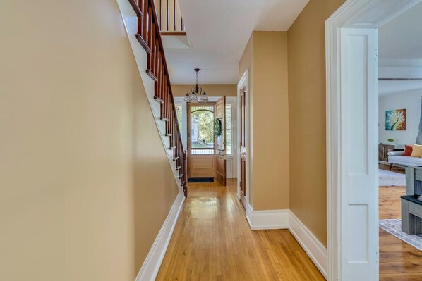 A hallway with wooden flooring, beige walls, and a staircase with wooden railings leading to an open door.