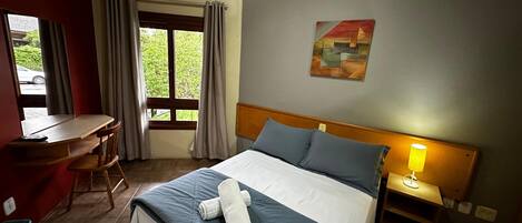 Una habitación de hotel con cama, escritorio, silla, ventana con cortinas, lámpara de noche y un cuadro en la pared.