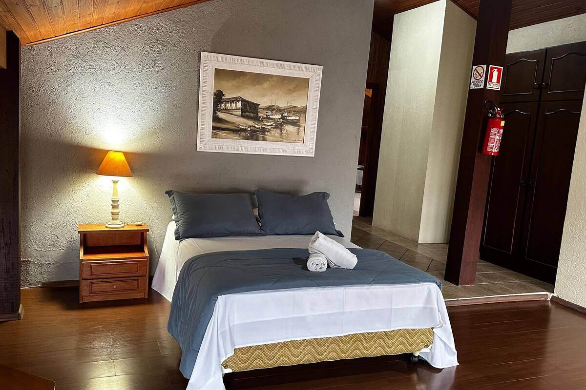 Une chambre à coucher avec un lit, une table de chevet avec une lampe, un tableau encadré au mur et un extincteur.