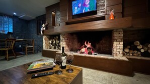 A cozy living room with a fireplace, a television, and a coffee table with books and wine glasses. - Pousada Canela Garden (Canela)