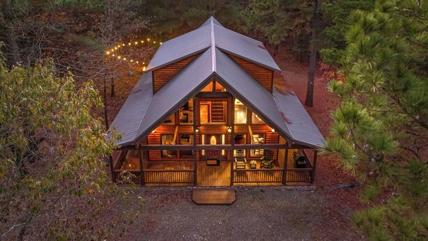 A cabin with a triangular roof, wooden exterior, and a porch with seating. - Beautiful cabin tucked away on almost 1.5 acres (Broken Bow)