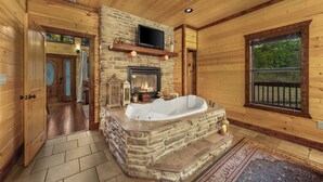 A log cabin interior with a stone fireplace, a large bathtub, and a television mounted above the fireplace. - Beautiful cabin tucked away on almost 1.5 acres (Broken Bow)