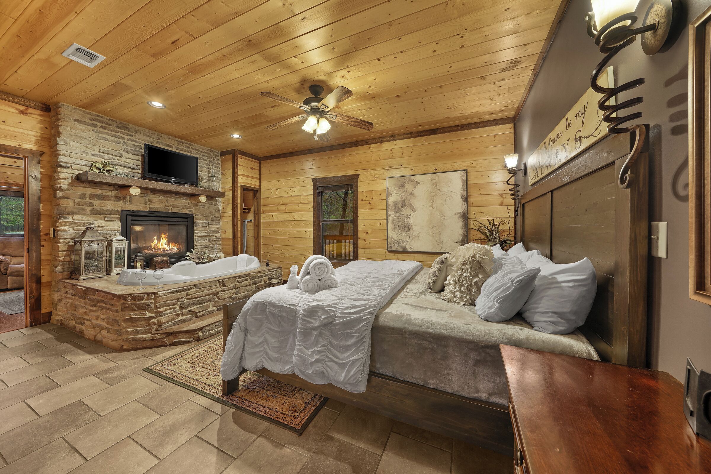 A log cabin interior with a stone fireplace, a bed with white bedding, a wooden ceiling, and a mounted television.