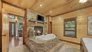 A log cabin interior with a stone fireplace, a large bathtub, and a television mounted above it. - Beautiful cabin tucked away on almost 1.5 acres (Broken Bow)