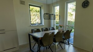A modern dining area with a wooden table and six chairs, large windows with a view of greenery, and a wall-mounted clock. - Holiday Home in Ewijk in a Beautiful Area (Ewijk)