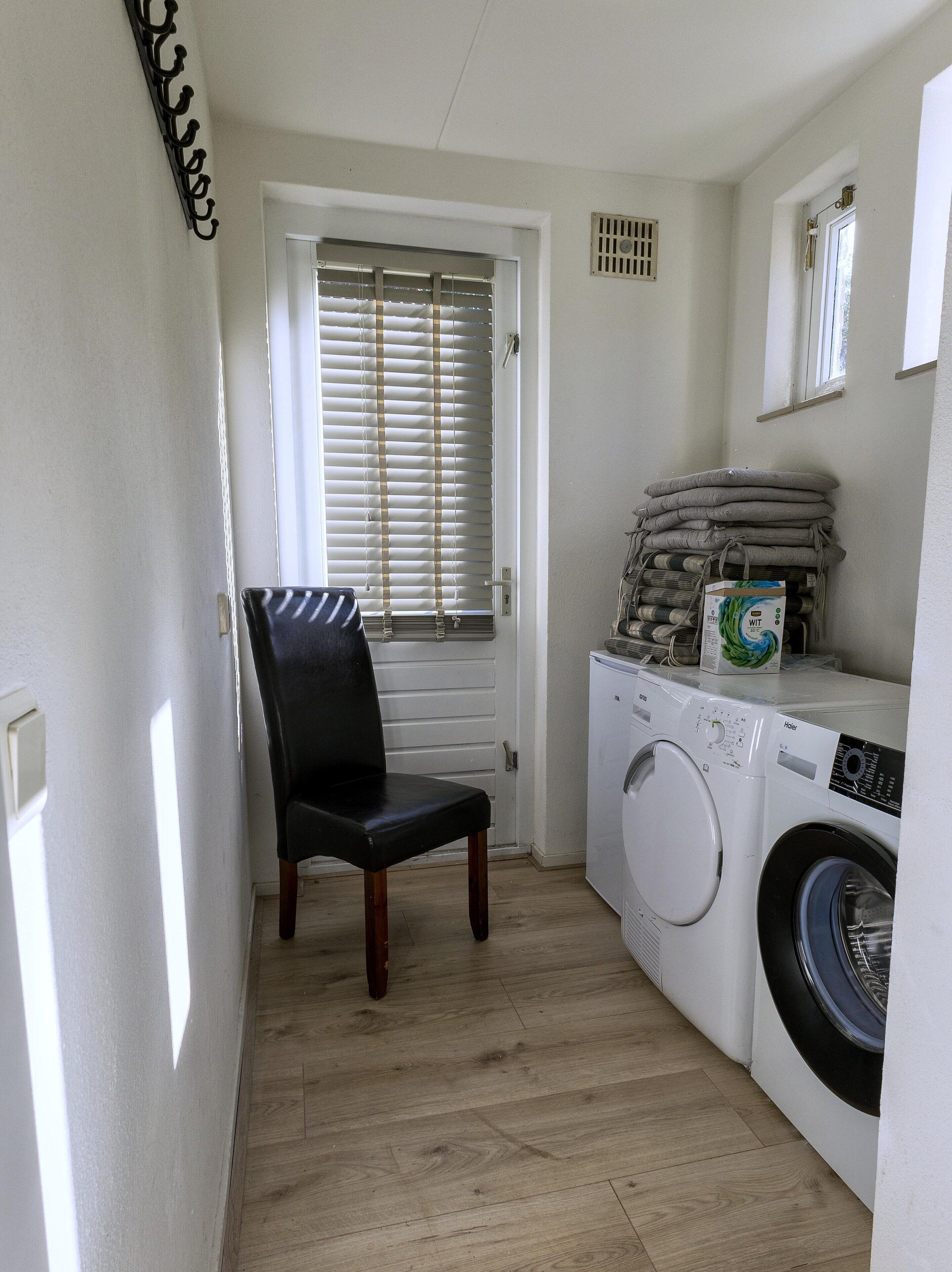 A laundry room with a chair, a washing machine, and a window with blinds.
