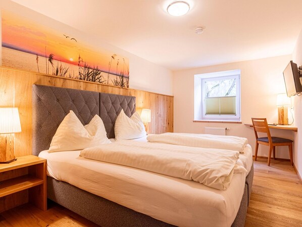 Cama doble con cabecera capitonada, dos mesitas de noche con lámparas, un escritorio de madera con silla y una ventana con persianas.