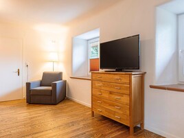 Una habitación con piso de madera, un sillón gris, un aparador de madera con cajones, un televisor encima y una ventana con persianas.