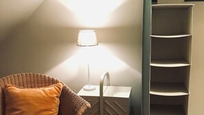 Et hjørne af et rum med en fletstol, et lille hvidt bord med en lampe og en reol.