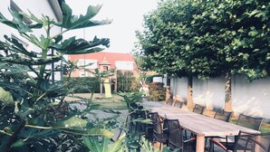 En udendørs terrasse med et træbord og stole, omgivet af grøn vegetation og en hvid væg.