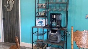 A dining area with a wooden table and chairs, a teal wall, and a metal shelf unit holding kitchenware and appliances.