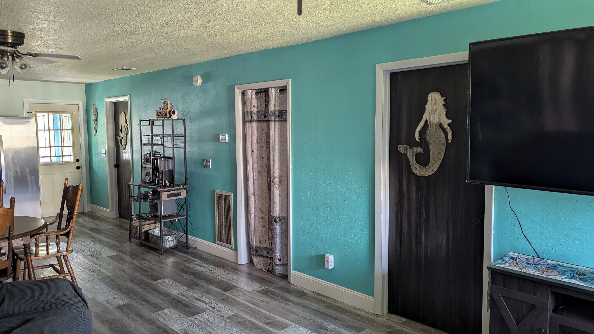 A room with teal walls, a wooden floor, a dining area with a table and chairs, a kitchen area with appliances, and a mounted TV.