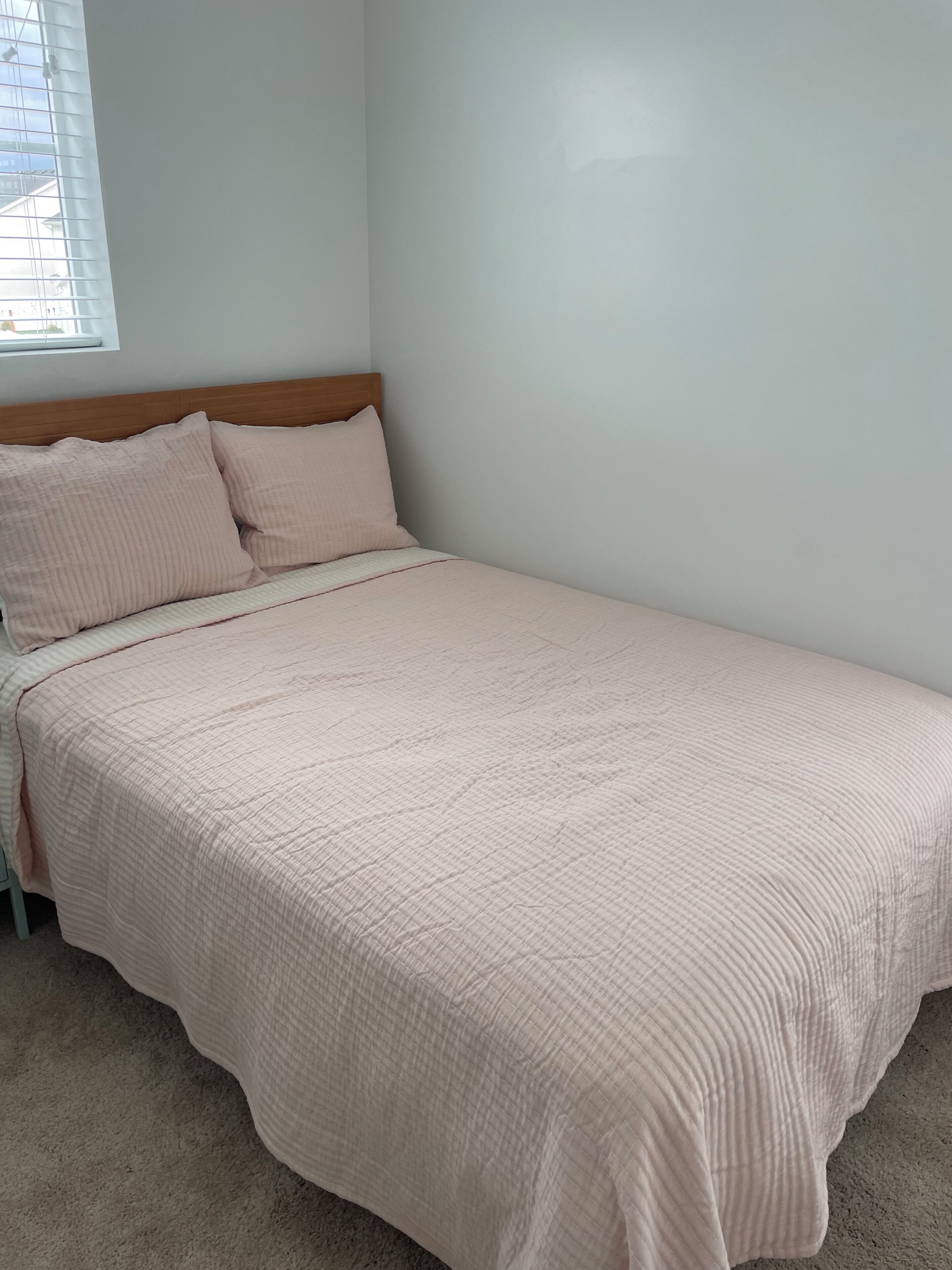 A single-bed room with a wooden headboard, a beige carpet, and a window with blinds.