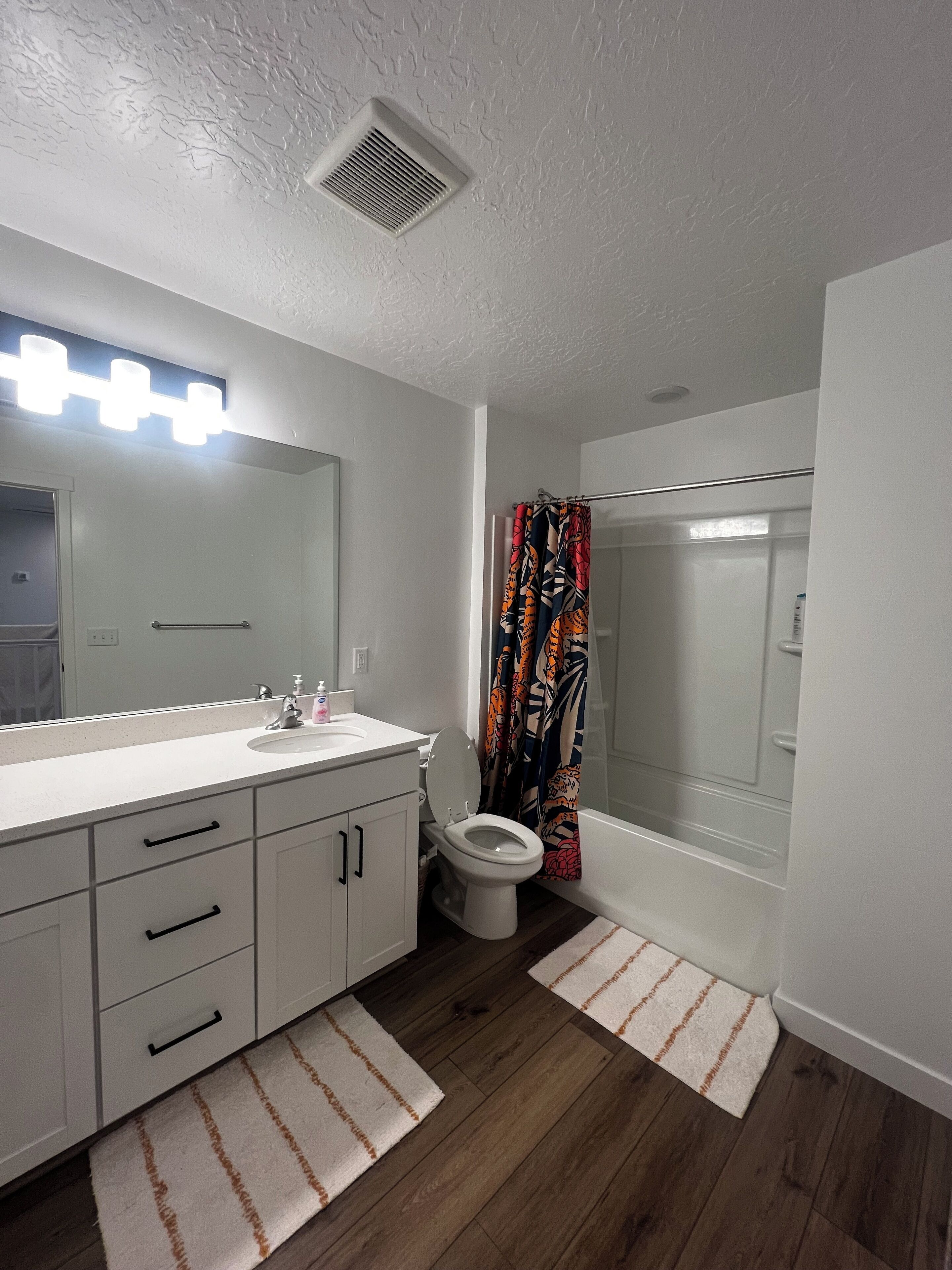 Un baño con tocador blanco, un espejo con luces, un inodoro y una ducha con cortina colorida.