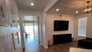A spacious living room with hardwood flooring, a flat-screen TV mounted on the wall, a dark-colored sofa, and a white rug. -  Beautiful home near Thanksgiving Point. Views of Utah Lake, sunrise and sunset (American Fork)