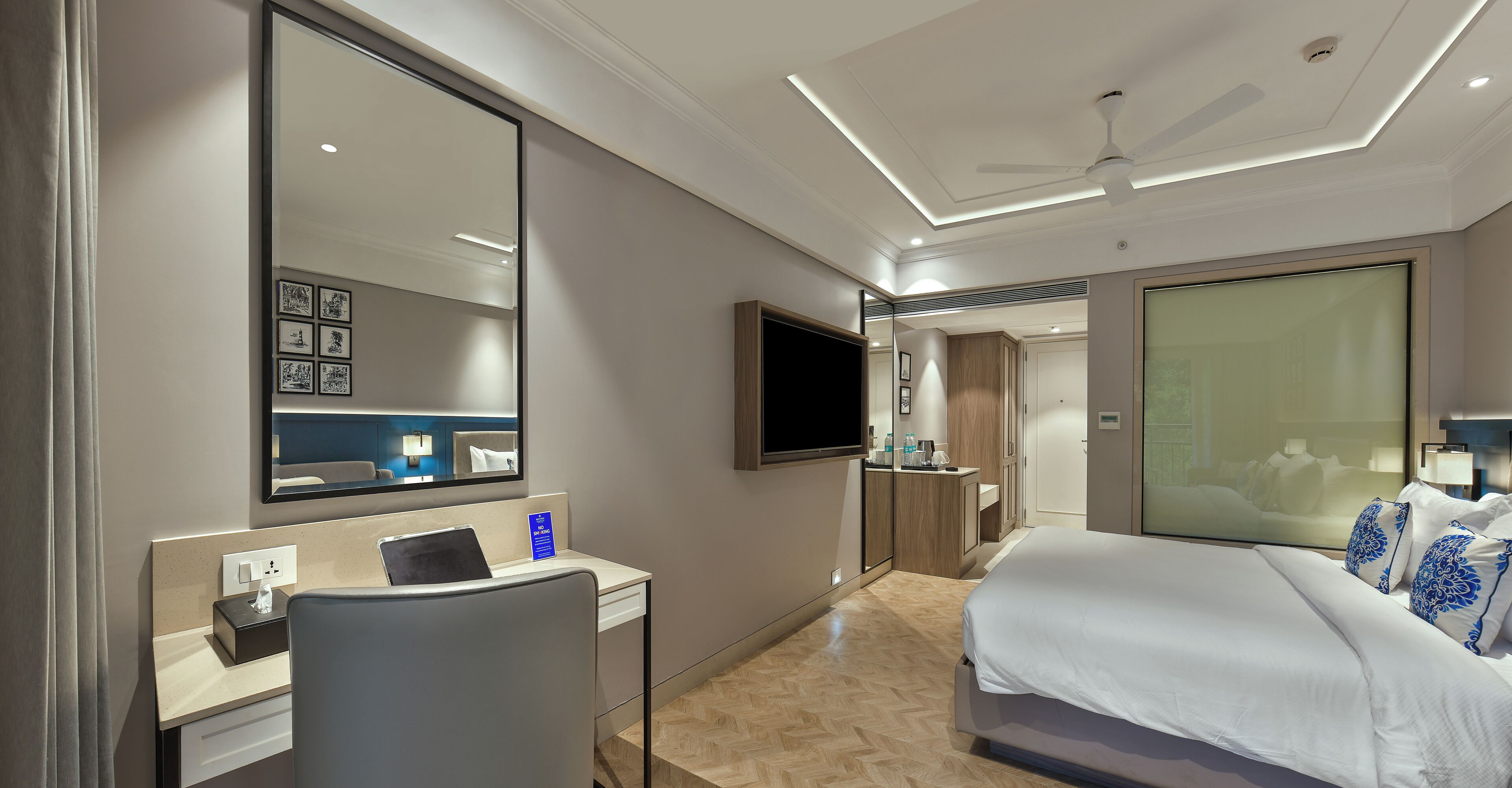 A modern hotel room with a large bed, a desk with a computer, a flat-screen TV, and a large mirror.