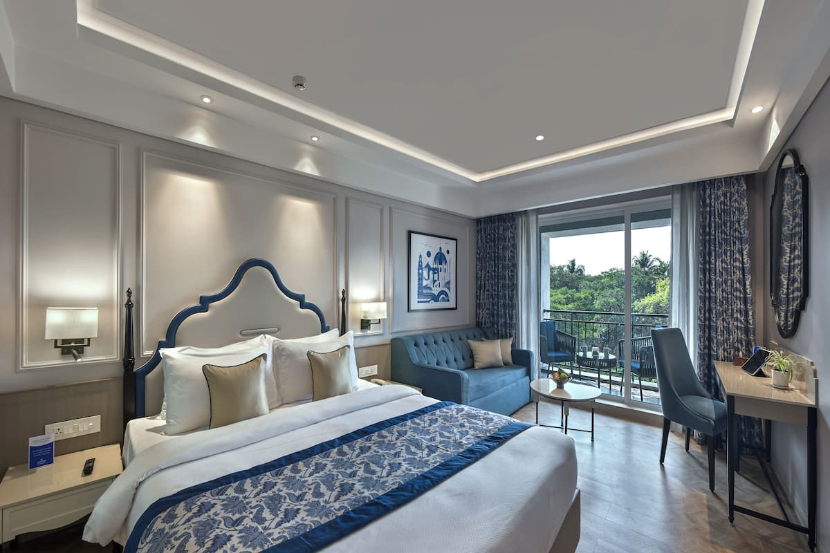 A hotel room with a large bed, a desk, a chair, a balcony with a view, and a framed picture on the wall.