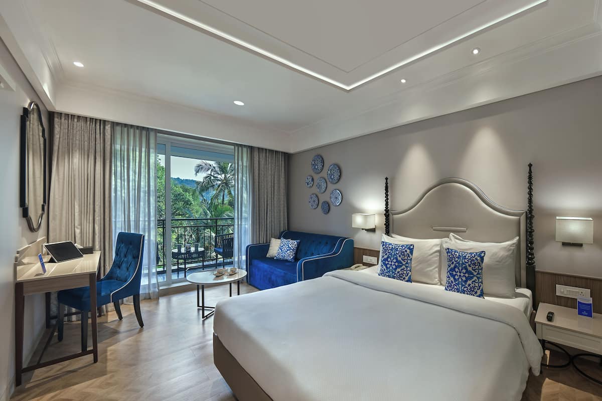 A hotel room with a large bed, a desk with a laptop, a blue armchair, a sofa, and a balcony with a view of greenery.