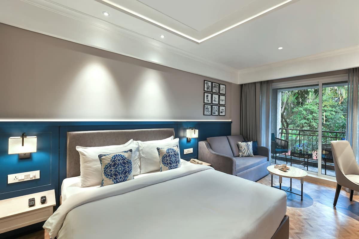 A modern hotel room with a large bed, a desk, a chair, and a balcony with a view of greenery.