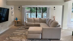 Ein modernes Wohnzimmer mit einem Ecksofa, einem Flachbildfernseher und einem großen Fenster mit Vorhängen.
