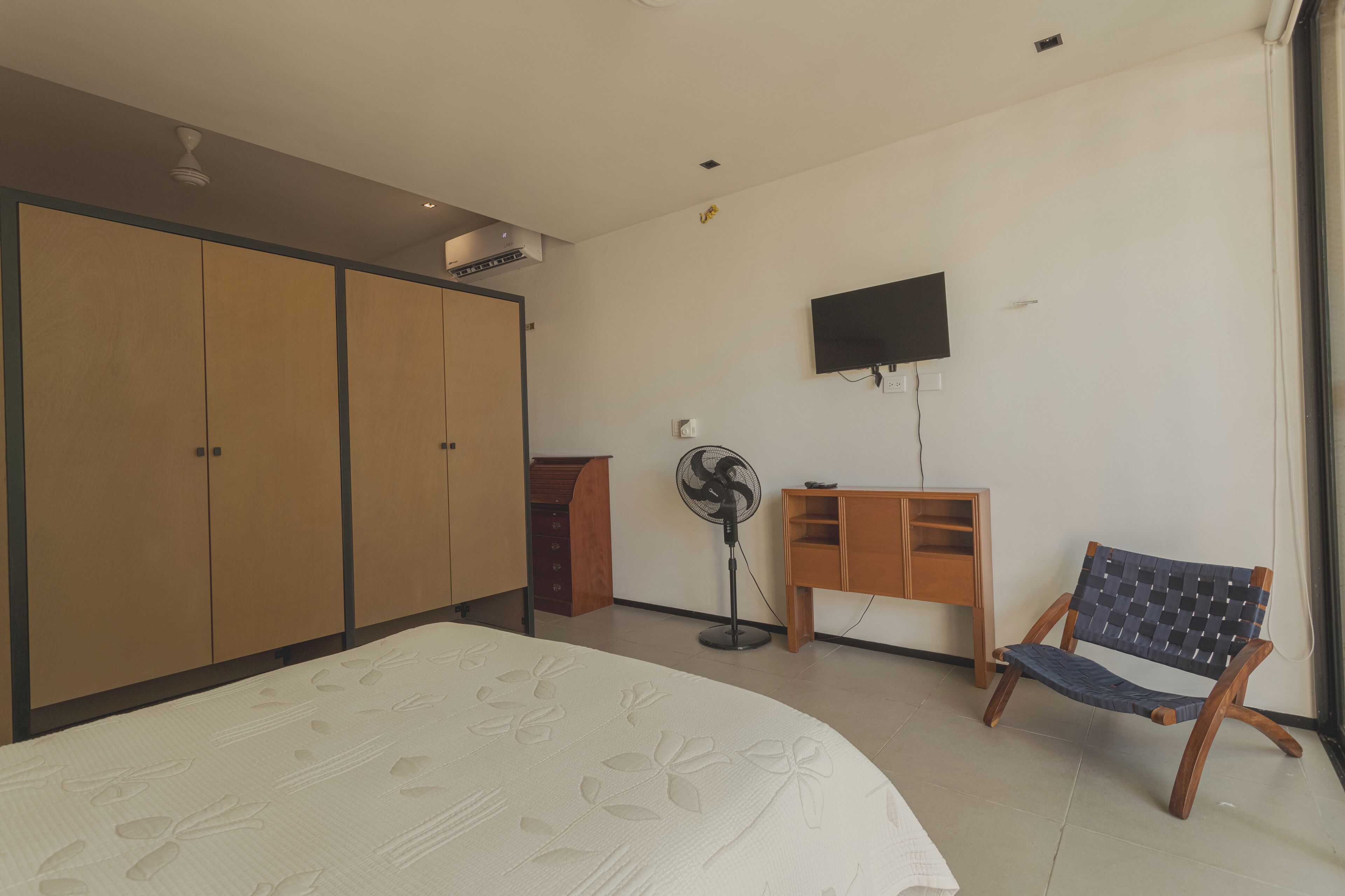 Un dormitorio con cama, armario, escritorio, silla, ventilador y televisor.
