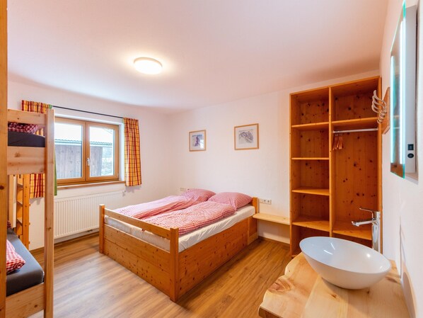 A bedroom with a wooden bunk bed, a single bed with checkered bedding, a large wooden wardrobe, a built-in wooden shelf, a white sink, and a window with curtains.