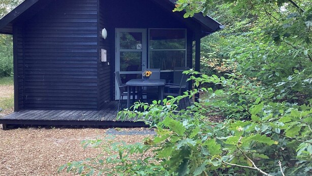 A small, dark wooden cabin with a porch and a table with chairs. - 6 pax Camping Cabin in Rodhus, Pandarup (Pandrup)