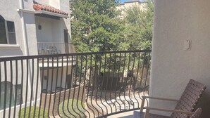 A balcony with two wicker chairs and a small table, a metal railing, and a view of a garden and buildings in the background.