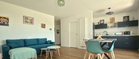 Een moderne woonkamer met een blauwe bank, een eettafel met stoelen, en een keuken met donkere kasten en witte apparaten.