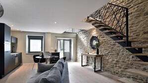 A modern living room with a grey sofa, a dining area, a kitchen, and a stone wall with a staircase. - Cocoon Rance - One Bedroom House, Sleeps 2 (Plouër-sur-Rance)