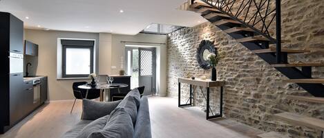 Un salon moderne avec un canapé gris, un coin repas, une cuisine et un mur en pierre agrémenté d’un escalier.