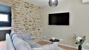 A living room with a stone accent wall, a grey sofa, a flat-screen TV, a small table with a vase of flowers, and a patterned rug. - Cocoon Rance - One Bedroom House, Sleeps 2 (Plouër-sur-Rance)
