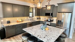 A modern kitchen with dark cabinets, a large island with stools, stainless steel appliances, and pendant lighting. - Fantastic New 8bd-4ba Betsie River Home, Game Rm, Two Family Rms, Kayaks, 6acres (Thompsonville)