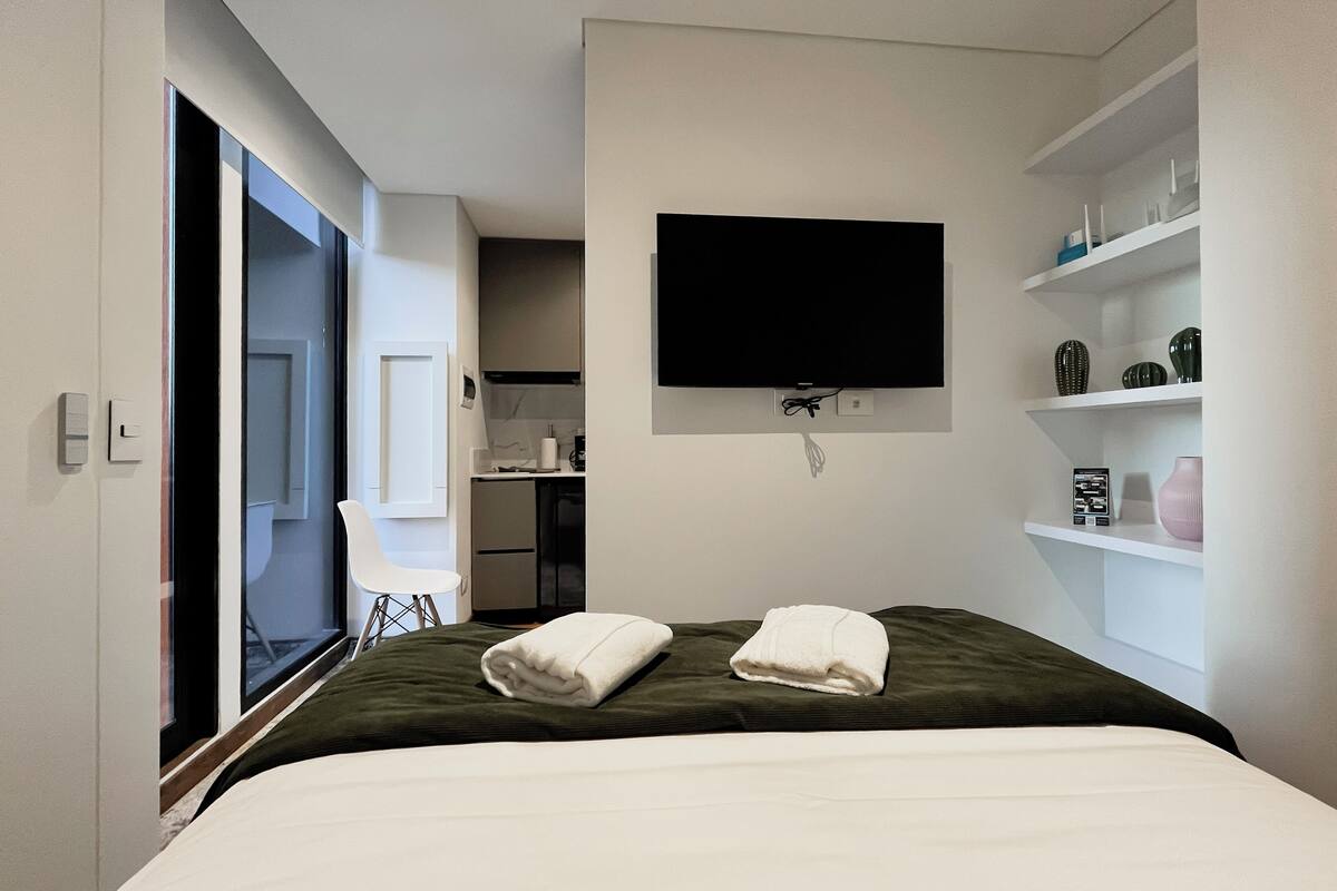 A modern hotel room with a bed, a television mounted on the wall, and a small kitchenette in the background.