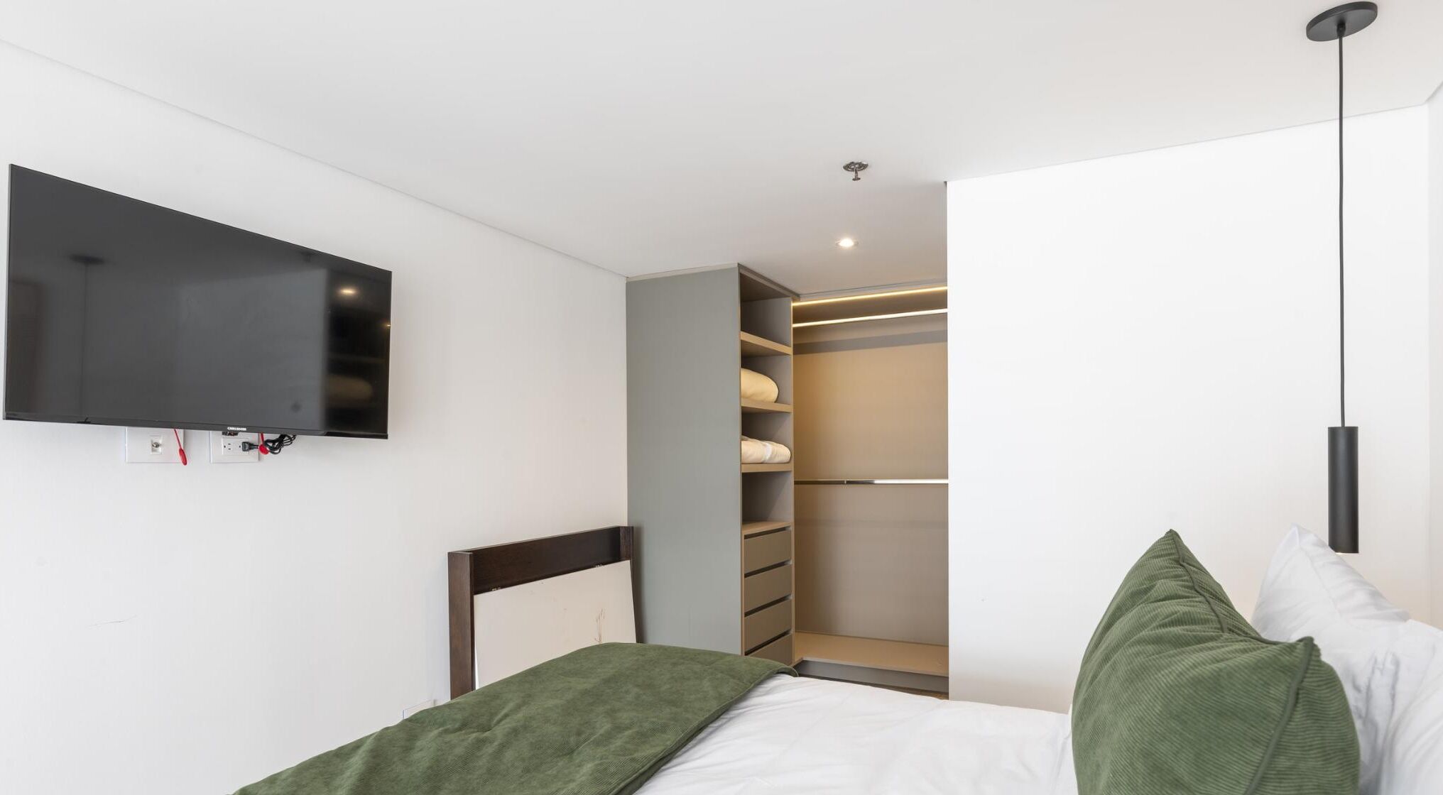 A bedroom with a bed, a TV mounted on the wall, a closet with shelves, and a hanging light fixture.