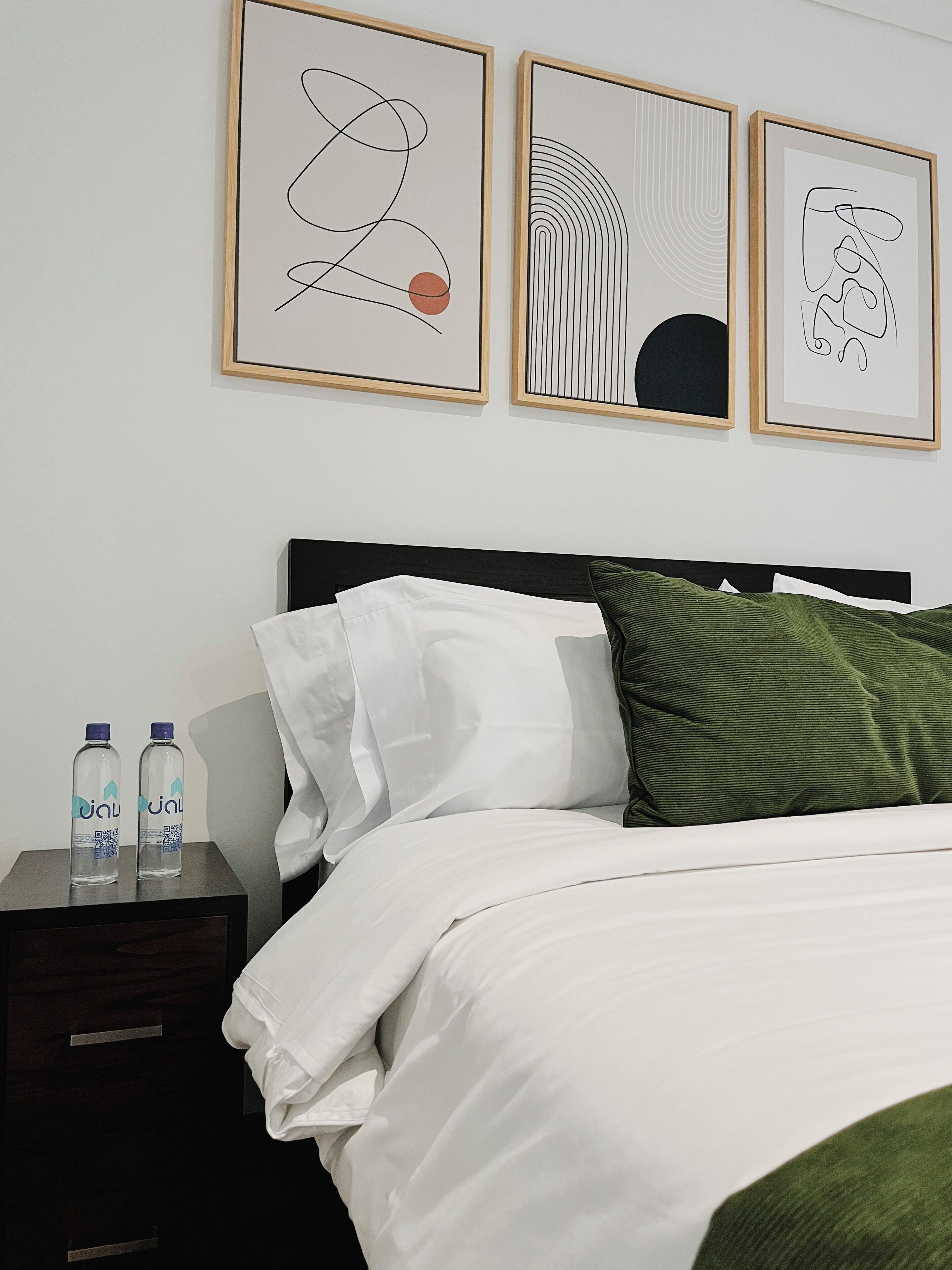 A neatly made bed with white and green pillows, a dark headboard, and two water bottles on a bedside table. Three abstract artworks are hung on the wall above the bed.