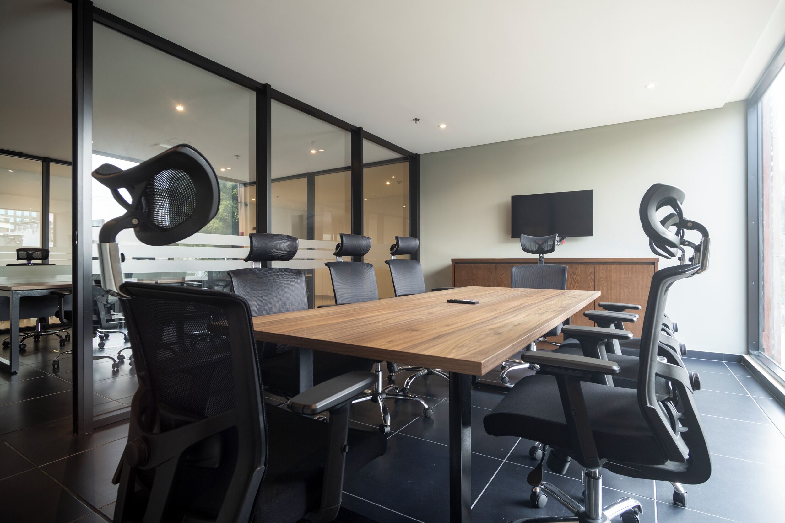 A conference room with a wooden table, black office chairs, a large window, and a flat-screen TV on the wall.