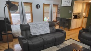 A living room with a black leather sofa, a black coffee table with a game, a floor lamp, and a view of a kitchen and dining area through a window. - Charming house with WiFi and AC in superb Randolph (Randolph)