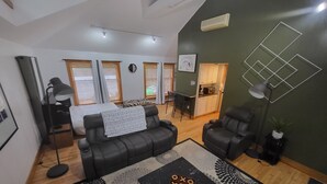 A modern living room with a black leather sofa, a coffee table, and a patterned rug. There is a kitchen area with a dining table and chairs visible through the windows. - Charming house with WiFi and AC in superb Randolph (Randolph)