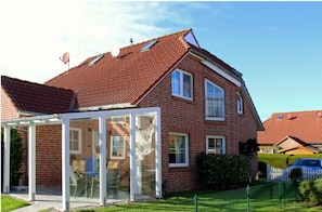 A two-story brick house with a red-tiled roof, a glass conservatory, and a white picket fence.