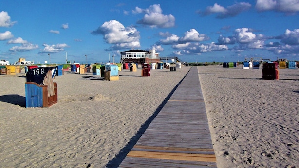 A beach with a wooden walkway leading to a building with a large window. There are colorful beach huts numbered 19, 193, 142, 466, 141, 174, 101, 121, 112, 185, and 143. - Haus in Nessmersiel am Wattenmeer (Dornum)