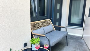Un sofá de mimbre con cojines, una mesa de vidrio con dos copas de vino y una planta en maceta en un patio con baldosas.