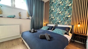 Un dormitorio con una cama grande, un radiador, una ventana con cortinas, una pared con estampado de hojas tropicales y una mesita de noche con una lámpara.