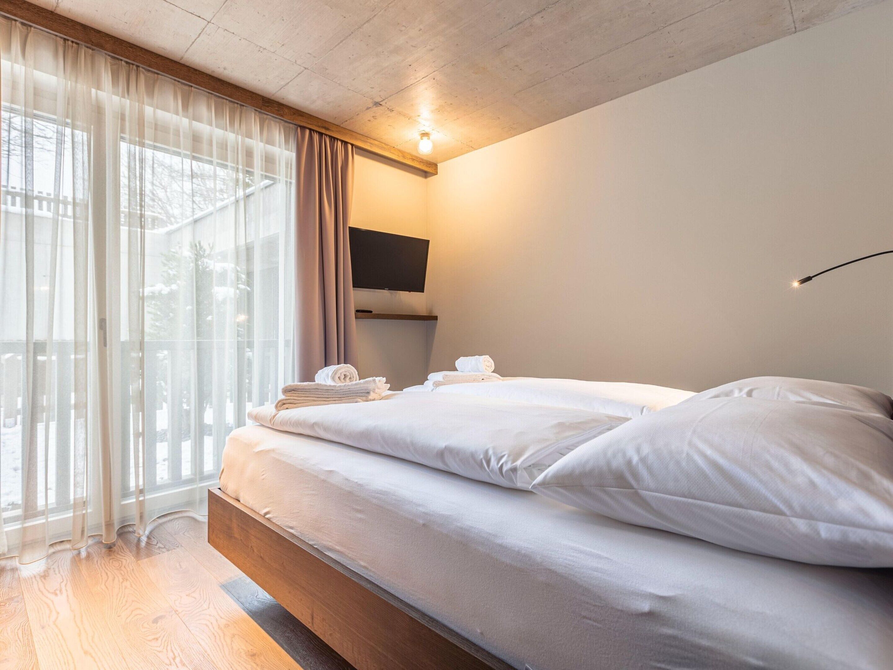 Ein modernes Hotelzimmer mit einem großen Bett, einem flachbildfernseher an der Wand und Blick ins Freie durch transparente Vorhänge.