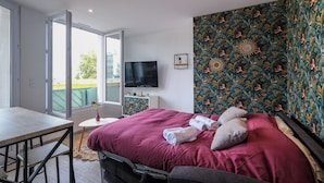 Un dormitorio con cama, televisión, una mesita y ventana.