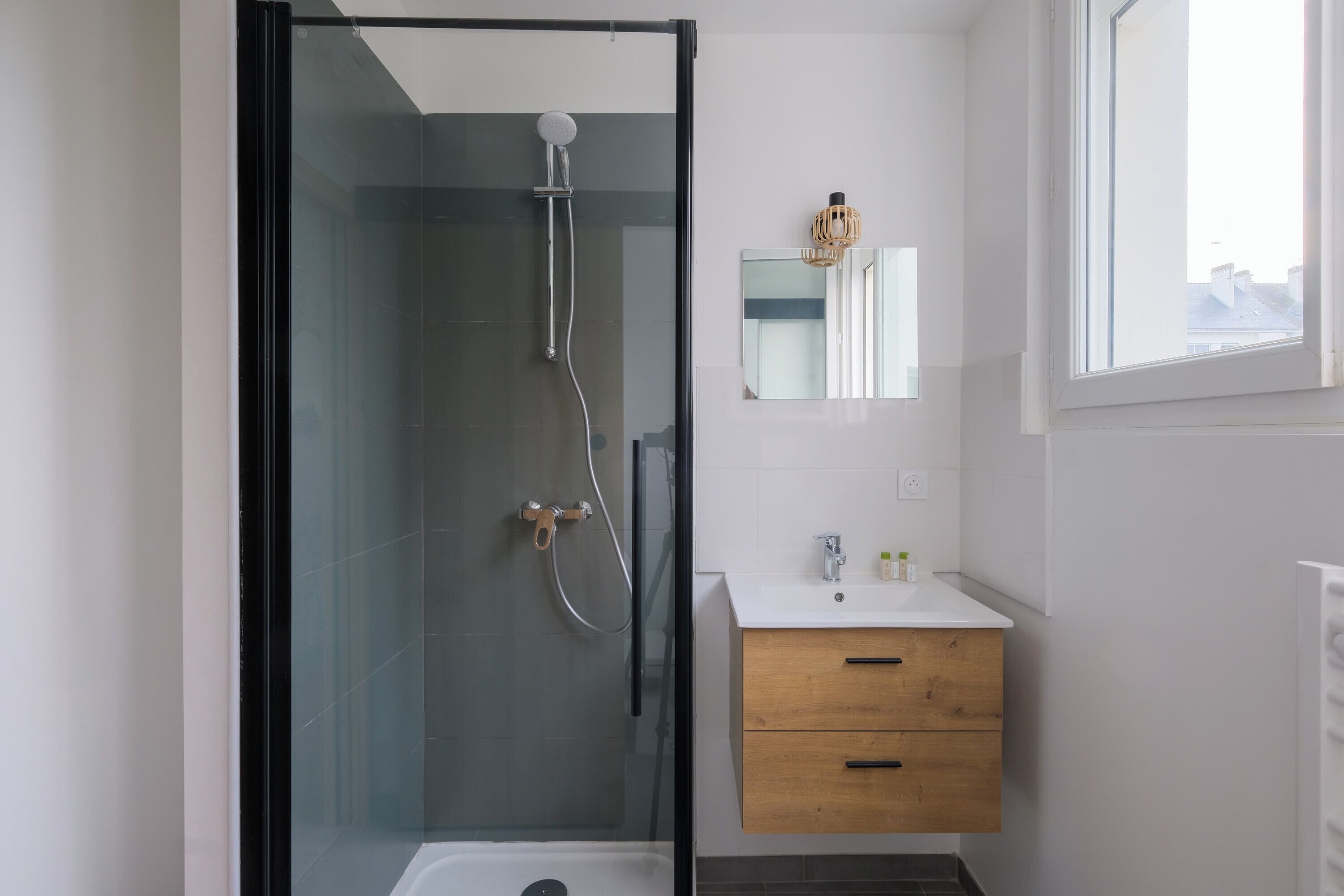 Een moderne badkamer met een douchecabine van glas, een houten wastafelmeubel met een spiegel, en een raam dat voor natuurlijk licht zorgt.