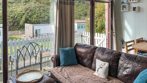 Un salon accueillant avec un canapé en cuir, un plateau en osier et une vue sur une clôture blanche et une végétation luxuriante à l’extérieur.