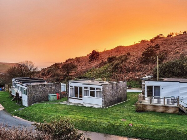 A row of mobile homes with stone facades, a grassy yard, and a hill in the background.
